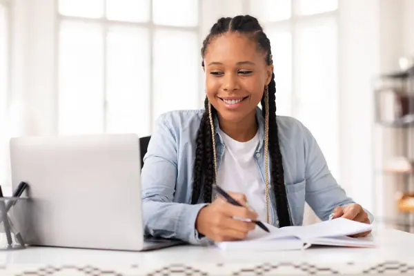 在家里用笔记本电脑学习 坐在明亮的房间里边上网边学习 用辫子写笔记边笑着年轻的黑人女学生 — 图库照片