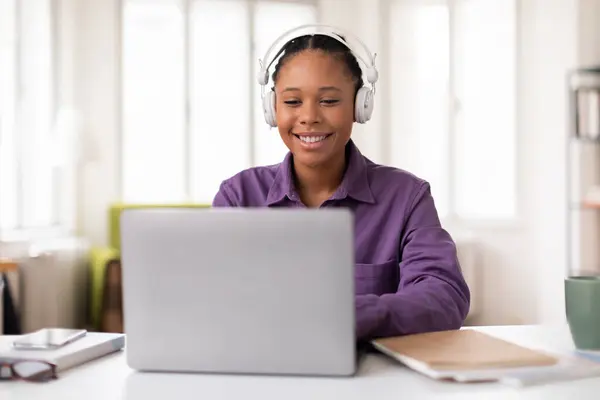 快乐的黑人女生 戴着辫子 头戴耳机 身穿紫色衬衫 在明亮的家庭写字台上的笔记本电脑上工作 对着屏幕微笑 — 图库照片