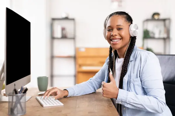 ヘッドフォンを身に着けている幸せな黒人女性学生は 自宅でオンライン教育を楽しんで コンピュータデスクでオンラインで勉強しながら親指を与えます — ストック写真