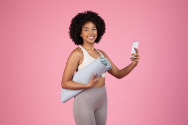 Işıl ışıl, spor kıyafetli, selfie 'ye gülümseyen, yoga minderini kolunun altında tutan ve canlı pembe arka planda cep telefonu kullanan Afro-Amerikan kadını.