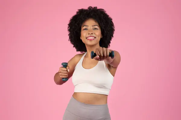 ラディアントの若い黒人女性は 重みを持ち上げながら笑顔を浮かべ ダンベルワークアウトに従事し ピンクのスタジオの背景にポーズ — ストック写真