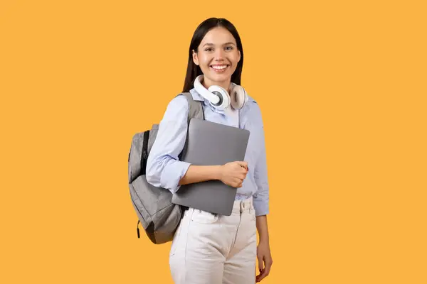 コンテンツと準備ができて学ぶ女性学生は 彼女の首の周りにヘッドフォンを着用しながらノートパソコンを保持し 黄色の背景に対する準備と現代の研究習慣を紹介しています — ストック写真