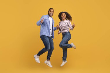 Enerjik genç siyah çift havada zıplıyor ve kameraya başparmaklarını doğrultuyor. Neşeli Afrika kökenli Amerikalı erkek ve kadın sarı stüdyo arka planında eğleniyor.