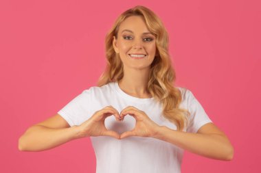 Aşk jesti. Gayri resmi beyaz tişörtlü güzel sarışın kadın parmaklarıyla bir kalp şekli oluşturuyor. Romantizmi ve minnettarlığı simgeliyor. Pembe stüdyo arka planında poz veriyor. Flört ve yardım kavramları
