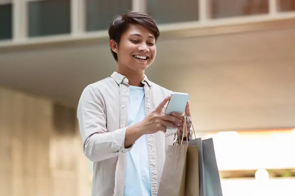 Happy young asian man with paper bags purchases in his hand using smartphone, checking promo on application enjoying shopping at city center. Cashback, e-commerce concept