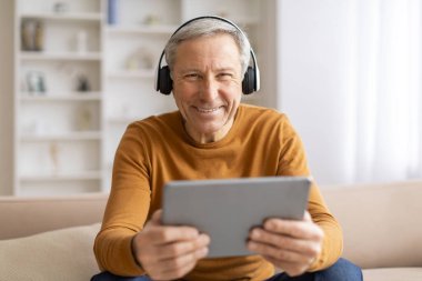 Evde dijital tablet ve kablosuz kulaklık kullanan mutlu gülümseyen yaşlı adam oturma odasında kanepede oturuyor, müzik dinliyor, güzel bir film izliyor. Dijital dünya ve yaşlılar