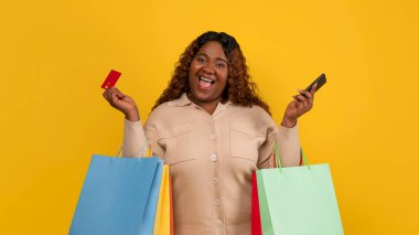 E-ticaret. Aşırı neşeli Afro-Amerikan tombul milenyum kadın internetten alışveriş yapıyor, elinde renkli kağıt torbaları, kredi kartı ve akıllı telefon var, sarı stüdyo arka planında izole edilmiş.