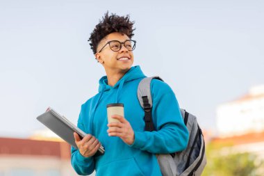 Akademik ara. Gülümseyen Brezilyalı öğrenci, elinde dizüstü bilgisayar ve kağıt kahve fincanı ile kampüs parkına bakan paket kahveyle gezmeyi seviyor. Portre çekimi