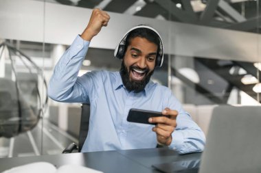 Heyecanlı milenyum sakallı Hintli eleman işyerinde video oyunu oynuyor, bilgisayarın başına oturuyor, akıllı telefon ve kablosuz kulaklık kullanıyor. Ofis çalışanları için stres atma