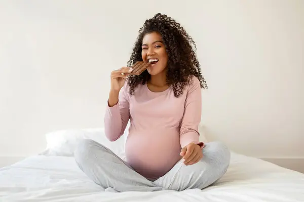 快乐的年轻黑人孕妇 在家里盘腿坐在白色的床上 享受着巧克力棒 快乐的非洲孕妇 在怀孕期间有着甜食的渴望 — 图库照片