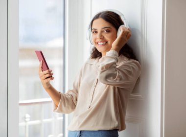 Kablosuz kulaklık takan mutlu genç bir kadın akıllı telefondan müzik dinlemenin keyfini çıkarıyor, güzel bir bayan gülümsüyor güneşli odada pencerenin kenarında duruyor, evde dinlenmenin keyfini çıkarıyor, bedava uzay.