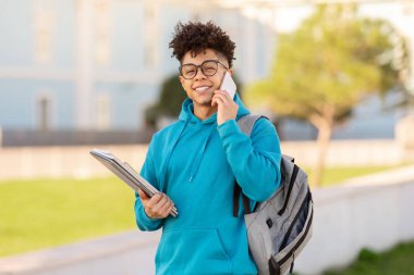 Sırt çantası ve dizüstü bilgisayarla cep telefonuyla konuşan gülümseyen siyahi üniversiteli çocuk, teneffüste iletişim kurarken kameraya bakıyor, kampüs parkında ayakta duruyor.