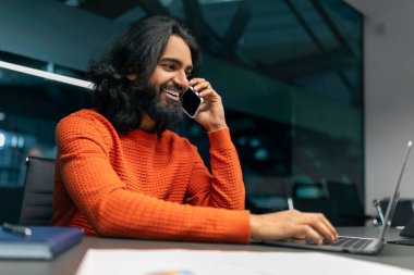 Hintli genç işadamı karanlık ofiste birden fazla iş yapıyor, masada oturuyor, dizüstü bilgisayarda klavye yazıyor ve asistanıyla telefon görüşmesi yapıyor, kopyalama alanı. Dijital dünya ve iş dünyası