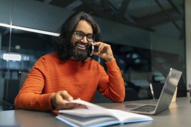Neşeli doğulu girişimci müşteriyle iş görüşmesi yapıyor, karanlık modern ofiste masa başında oturuyor, telefonla konuşuyor ve ajandada ajandaya bakıyor, kopyalama alanını kontrol ediyor. Zaman yönetimi