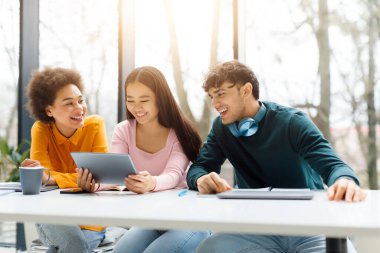 Neşeli genç öğrenciler tablet paylaşıyor ve kahkaha ve üretkenlikle dolu güneşli odada birlikte ders çalışırken kahveyle ve kahveyle eğleniyorlar.