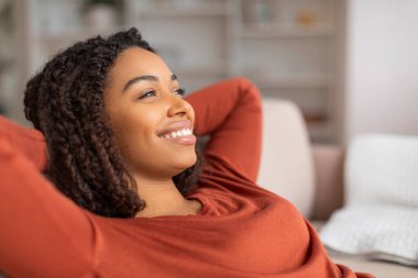 Güzel, gülümseyen siyah bir kadının portresi rahat koltuğa yaslanıyor, mutlu, sakin, Afro-Amerikan bir kadının elleri başının arkasında dinleniyor, evdeki oturma odasında dinleniyor,