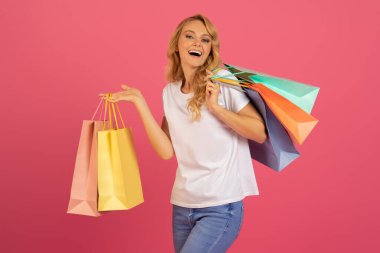 Mevsimlik Alışverişçi. Güzel Avrupalı sarışın bayan alışveriş torbaları taşıyor ve büyük satışlardan dolayı mutluluğunu ifade ediyor, stüdyo ortamında pembe arka planlı alışverişlerle ayakta duruyor. İndirim tanıtımı