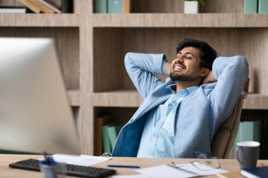 Rahat Hintli işadamı arkasını yaslıyor ve ellerini başının arkasına koyuyor, modern ofis iç bilgisayarının yanında otururken işe ara veriyor.