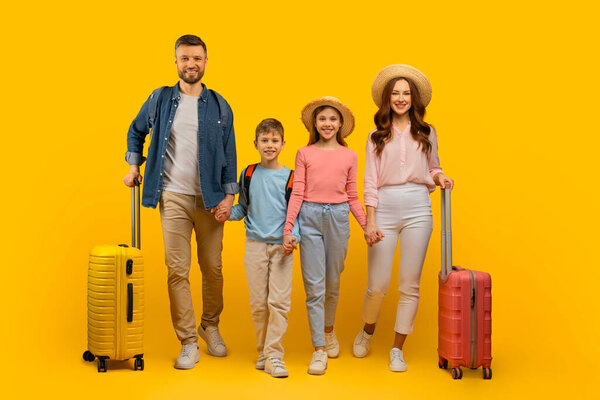 Happy Young Family Of Four Holding Hands And Walking With Suitcases On Yellow Studio Background, Cheerful Parents And Their Two Kids Wearing Straw Hats, Ready For Vacation Trip, Full Length Shot