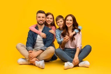 Dört kişilik mutlu, genç bir aile yerde oturuyor ve mutlu, sevgi dolu ebeveynler sarı stüdyo arka planında oğulları ve kızlarıyla birlikte poz veriyor, kameraya bakıyor, mekanı kopyalıyor.