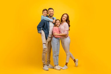 Aile portresi. Mutlu genç ebeveynler ve iki çocuğu sarı arka planda kucaklaşıyorlar, neşeli anne, baba, oğul ve kızın stüdyoda kucaklaşması, boşluğu kopyalaması...