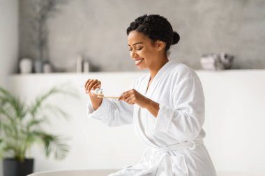 Bornozlu mutlu siyah kadın dişlerini fırçalamaya hazırlanıyor, diş macununu fırçaya sürüyor, gülümseyen Afro-Amerikan kadın evdeki şık minimalist banyoda günlük diş hijyeni yapıyor.