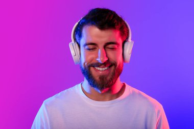 Bin yıllık kablosuz kulaklıklı adamın portresi kapalı gözlerle müzik dinlemeyi, dijital kulaklık kullanmanın sevincini, mor ve mavi neon ışıklarla aydınlatılmış stüdyo arka planında poz vermeyi seviyor.