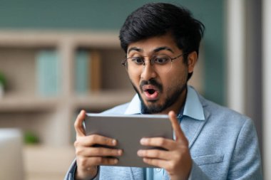 Heyecanlı Hindu girişimci, modern iş yerinde dijital tablet kullanıyor, duygusal iş adamının ofis içi bilgisayar aygıtında web sörfünün portre resmi.