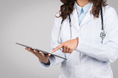 Dijital tabletle uğraşan, tıbbi bilgilere yön veren ya da giriş yapan kadın doktor, gri arka planda sağlık hizmetlerine teknolojik entegrasyonu gösteriyor