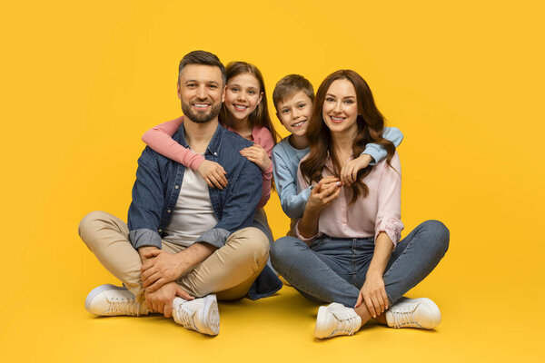 Happy young family of four sitting on floor and embracing, cheerful loving parents posing together with their son and daughter on yellow studio background, looking at camera, copy space
