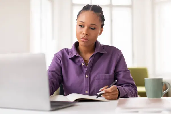 穿着紫色衬衫的专注的非洲裔美国女学生在笔记本电脑上记笔记 周围是温馨的家庭学习环境 还有一杯热茶 — 图库照片