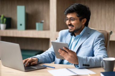 Bilgisayarında ve dijital tablet bilgisayarlarında çalışan Orta Doğulu bir yönetici modern ofiste masa başında oturuyor, kapalı alanda web sörfü yapıyor. Dijital iş ve yenilik, modern teknoloji