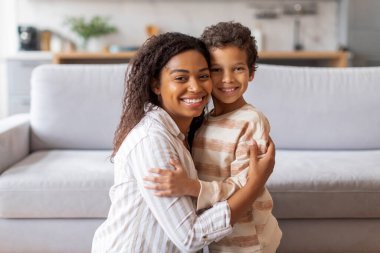 Neşeli siyah anne ve küçük oğlu evlerinde sıcak, sevgi dolu bir kucaklaşmayı paylaşıyorlar, Afrikalı Amerikalı anne ve erkek çocuk kameraya sarılıyor ve mutlu bir şekilde gülümsüyor, parlak, modern oturma odasında poz veriyorlar.
