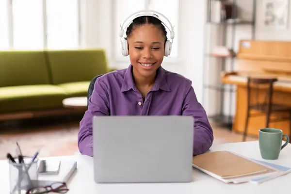 身穿紫色衬衫的黑人女学生穿着网络课程 戴着白色耳机 使用笔记本电脑 桌上放着笔记本电脑和杯子 微笑着 — 图库照片