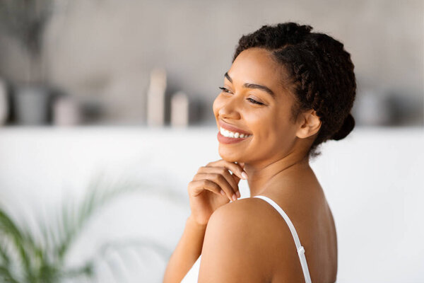 Portrait of happy young black woman wearing white tank top smiling and touching chin, charming african american female posing in bathroom interior, enjoying making beauty routine at home, free space