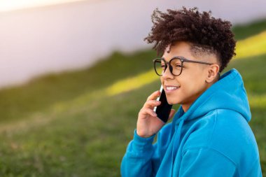 Akıllı telefondan sohbet eden neşeli Brezilyalı genç adamın yan görünüm portresi, dışarıda gözlüklerle poz veren, hoş bir telefon konuşması yaparken gülümseyerek başka yere bakan. Boş boşluk