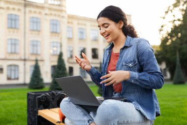 Doğu 'lu genç bir kadın bilgisayar aracılığıyla bilgisayar konferansı veren bilgisayar kamerasıyla konuşan öğrenci videosu dışarıda bankta oturarak öğreniyor. Modern Uzaklık Çalışması, E-Öğrenme Konsepti