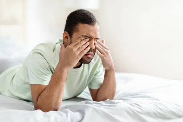Depression, mental disorder. Closeup of upset young black man laying on bed, rubbing his eyes. Unhappy millennial african american man crying, grieving, copy space