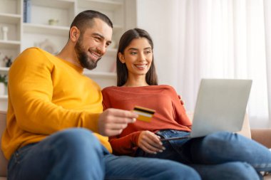 Kolay ödemeler. Laptop bilgisayarı ve kredi kartı olan mutlu genç çift evdeki koltukta dinleniyor, neşeli eşleri online alışveriş yapıyor, İnternet alışverişlerinin tadını çıkarıyor,