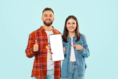 Bu uygulamayı seviyoruz. Kafkasyalı genç çift, Büyük Cep Telefonu 'nu Boş Ekran ile Gösteriyor, Mavi Stüdyo Arkaplanı Üzerinde Başparmak İşaretleri Yapıyor. Model, Kolaj