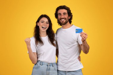 Neşeli mutlu Avrupalı genç kadın ve sakallı adam, elinde mavi bir kredi kartı olan ve her ikisi de beyaz tişörtlü, parlak sarı arka planda, stüdyo