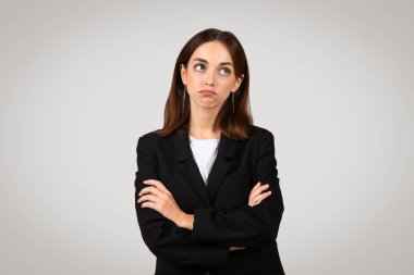 Kollarını kavuşturmuş siyah takım elbiseli, gri bir arka plan, stüdyo karşısında somurtkan ve şüpheli bir yüz ifadesi gösteren, memnun olmayan beyaz, profesyonel bir kadın.