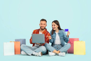 Neşeli genç çift kredi kartı ve dizüstü bilgisayar kullanıyor, mavi arkaplanda alışveriş çantaları ile oturuyor, Webstore 'dan kıyafet satın alıyor, karısı bilgisayar seçerken parmağını işaret ediyor.