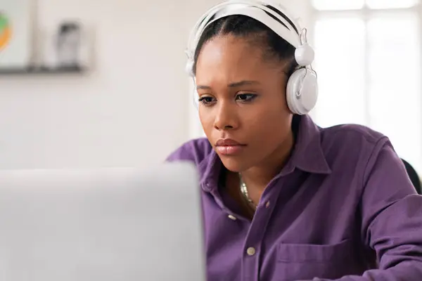 集中した黒人女性の学生は 彼女のラップトップスクリーンに集中し 白いヘッドフォンを身に着けている間 彼女の研究に魅了され おそらくオンラインで学ぶ — ストック写真