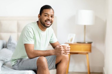 Mutlu, yakışıklı, sakallı, binlerce yıllık Afrikalı Amerikalı adamın evde pijama giymiş, elinde kupa tutarak kameraya gülümseyen, sabah kahvesini içen ve uzay fotokopisi çeken bir fotoğrafı.