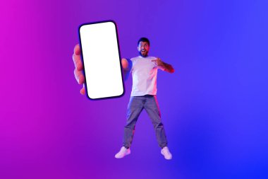 Stüdyoda genç sakallı bir adamın büyük boş bir ekranı işaret ederken neon mavi ışıkta atlayıp yeni uygulamanın heyecanını ifade ettiği bir fotoğraf. Modern teknoloji ve mobil iletişim