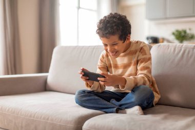 Neşeli küçük zenci çocuk, evinde rahat bir şekilde koltukta otururken akıllı telefonuyla oyun oynamaktan zevk alıyor. Elinde cep telefonuyla gülüyor, rahat bir oturma odasında dinleniyor.