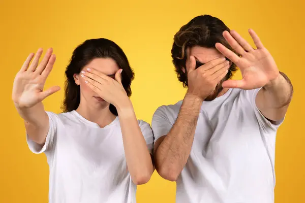 A man and woman in white shirts covering their eyes with hands, showing the see no evil concept, with a gesture of avoidance or not wanting to look, against a yellow background
