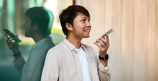 Side view of young asian guy standing next to building glass wall outdoors, recording audio message or task for AI assistant on smartphone, panorama with copy space