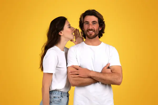 Confident happy caucasian man with crossed arms smiling while a woman whispers in to his ear, both dressed in white shirts against a vivid yellow background, studio. Gossip, news